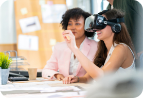 Two women sitting at a desk while the woman on the left has on virtual reality goggles and is drawing in the air at the same time.